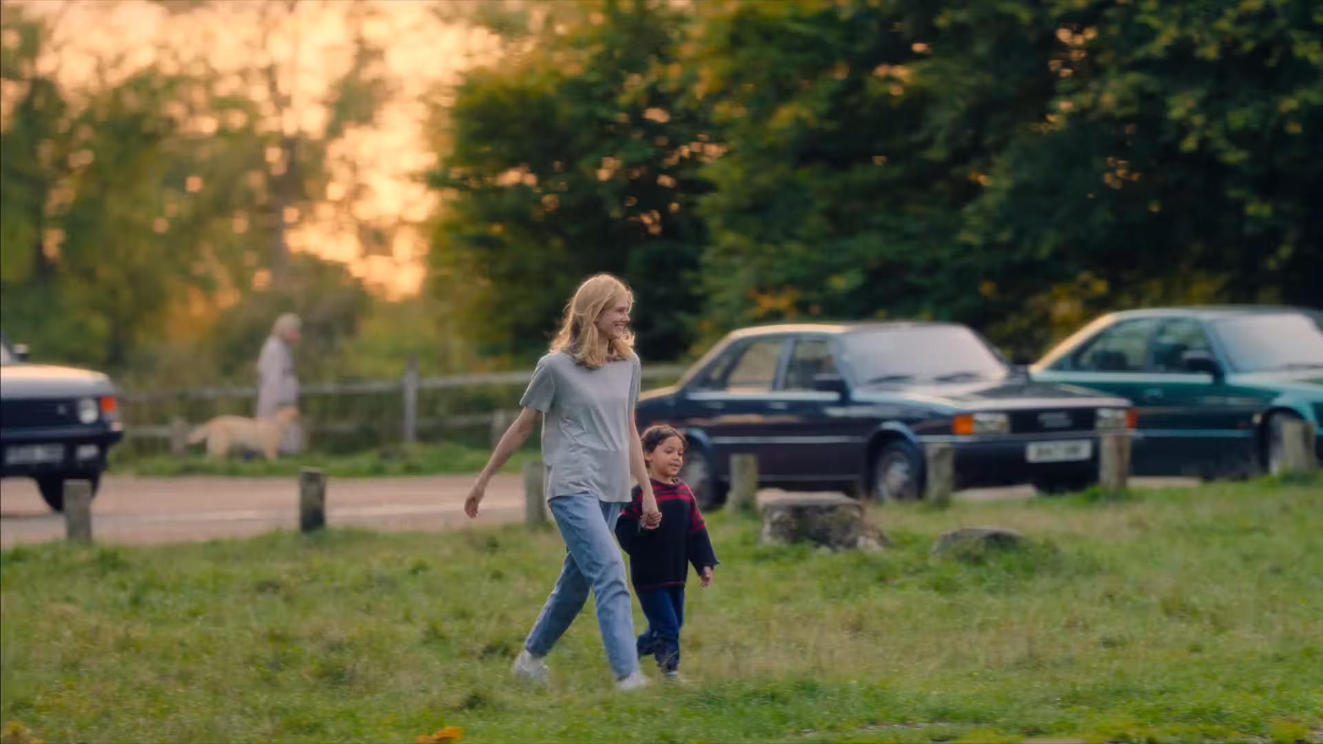 Woman walking through a park with her child, with parked cars visible in the background.